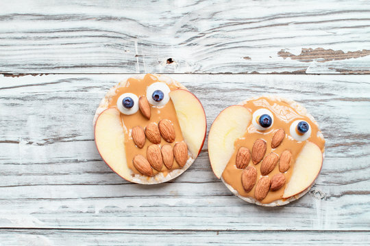 Funny Food For Kids. Rice Cakes In The Shape Of Owls Lying On A Rustic White Table. Owl Is Made From Peanut Butter, Apple Slices, Almonds, Candy And Blueberries. Top View, Flay Lay Position.