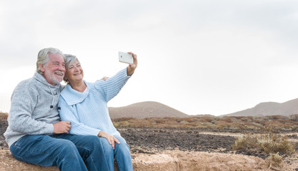 Two smiling old people. Senior couple with grey hair sitting on the cliff looking the mobile phone for a selfie. Clear sky. Waiting for the sunrise