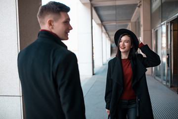 young man in a black coat goes towards a young woman in a black hat and coat
