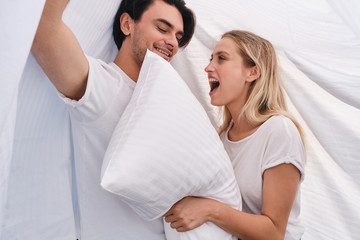 Handsome brunette man and pretty cheerful blond woman with white pillow in hands happily sitting and covering with blanket in bed at cozy home