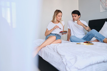 Thoughtful brunette man drinking coffee while pretty blond woman near eating pancakes with berries. Young attractive couple in white T-shirts having delicious breakfast in bed in modern hotel