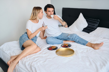 Obraz na płótnie Canvas Young brunette man and pretty blond woman dreamily drinking coffee together. Beautiful couple in white T-shirts having delicious breakfast in bed in modern cozy hotel