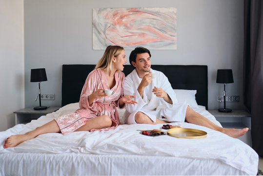 Handsome brunette man in white bathrobe and beautiful blond woman in pink velevt robe happily talking with cups of coffee in hands. Young couple having delicious breakfast in bed in hotel