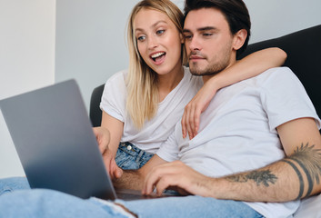 Young thoughtful brunette man and pretty emotional blond woman in white T-shirts using laptop together in bed at home