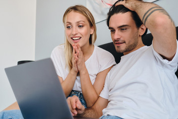 Young brunette man and pretty smiling blond woman in white T-shirt joyfully using laptop together sitting on bed in modern hotel