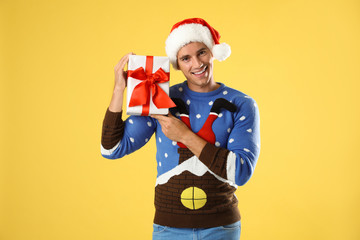 Happy man in Christmas sweater and Santa hat holding gift box on yellow background