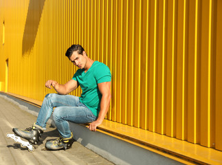 Handsome young man with inline roller skates sitting near yellow building, space for text