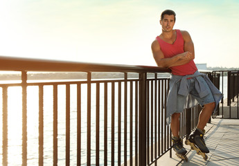 Handsome young man with roller skates on pier near river, space for text