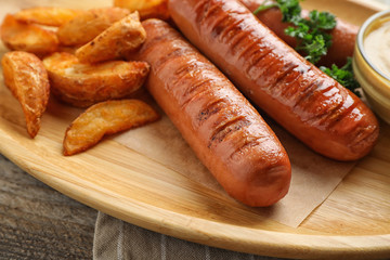Delicious grilled sausages and fried potatoes on wooden plate, closeup. Barbecue food