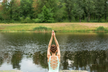 Fototapeta na wymiar Thin brunette girl plays sports and performs beautiful and sophisticated yoga poses in a summer park. Green lush forest and the river on the background. Woman doing exercises on a yoga mat