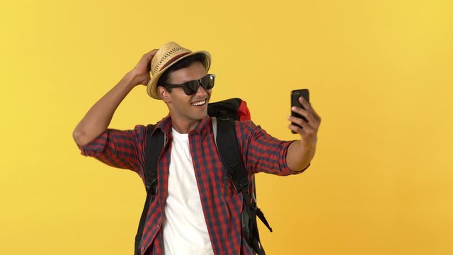 Young happy Indian tourist man with backpack smiling and waving bye while having video call on mobile phone against yellow background