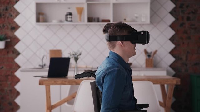 Disabled man sitting in comfort wheel chair and spending free time at room with natural soft light inside modern interior house. Male having fun using virtual reality helmet or glasses