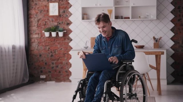 Adult disabled man sitting in special comfort wheelchair and spending day at home with bright cozy room with modern interior. Male wearing in denim jeans clothes using personal computer and smiling