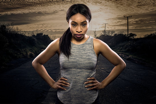 Athletic black African American female runner or jogger resting to pose with a motivated facial expression with a road during sunset in the background.  Depicts endurance and determination in marathon