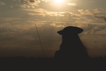 Silhouette of pirate woman in a pirate hat and with a sword in hands over a evening cloudy sky...
