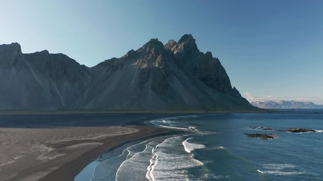 Aerial panning video of Stokksnes & Vestrahorn. Waves rolling in on black beach with mountain in background.