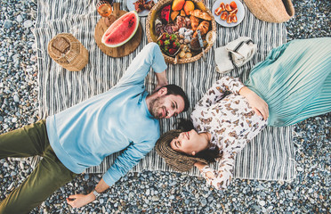 Summer beach picnic at sunset. Young couple lying on striped blanket, looking at each other at...