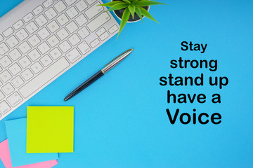 Flat lay, top view office table desk. Workspace with STAY STRONG STAND UP HAVE A VOICE inscription written with keyboard, pen and sticky notes on blue background. Business, Insurance and motivation co