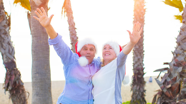 Happy excited senior couple celebrating winter holidays on the beach wearing Christmas Santa's red hats - smiling retirees people hugging and raising hands for xmas