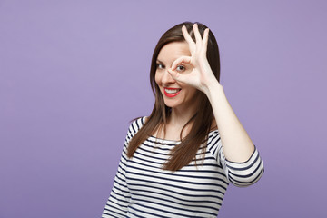 Pretty young woman in casual striped clothes posing isolated on violet purple background in studio. People lifestyle concept. Mock up copy space. Holding hand near eye imitating glasses or binoculars.