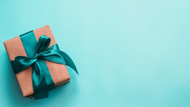 Gift box in craft wrapping paper and green satin ribbon on turquoise blue background, copy space right. Beautiful Christmas, New Year or Birthday present, flat lay or top view. Banner