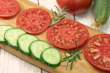 Red tomato and green cucumber lie on the board. Summer fresh vegetables cut into slices. Sprigs of rosemary decorate.