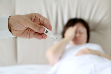 Ill woman laying in the bed, digital thermometer in male hand. Doctor measuring body temperature, concept of cold and flu, fever, illness