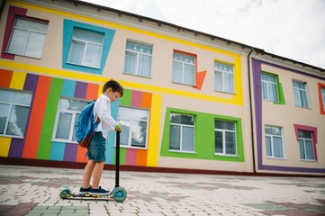 Teenage boy with kick scooter near modern school. Child with backpack and book outdoors. Beginning of lessons. First day of fall. Back to school.