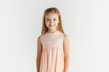 Stylish little smiling girl posing in dress isolated on white studio background. Caucasian blonde female model. Human emotions, facial expression, childhood. Looks calm, standing, looking at camera.