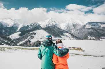 Back view photo of young loving couple hugging over winter mountains. Looking at mountains.