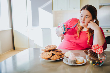 Fat young woman in kitchen sitting and eating sweet food. Bored plus size model hold blue soft tape measure in hand and look at it. Body positive. Cookies and pancakes on table. Daylight in kitchen.