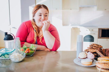 Fat young woman in kitchen sitting and eating food. Looking at sweet junk meal on left side. Temptation. Healthy food on left side. Hard choice. Body positive.