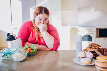 Fat young woman in kitchen sitting and eating food. Looking at sweets on right side. Salad and other healthy food on left one. Temptation. Body positive.