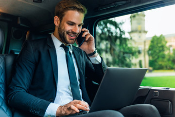 Handsome young man in full suit talking on smart phone and working on laptop, discussing job questions and smiling while sitting in the car