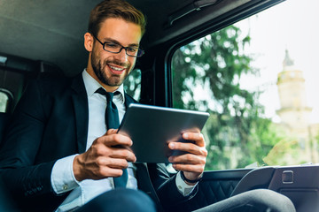 Smiling young man in full suit working using digital tablet while sitting in the car