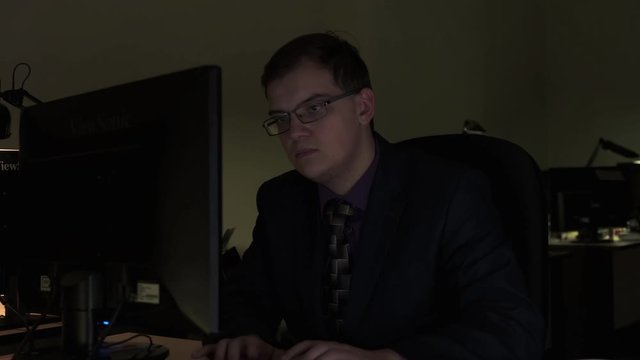 Male office worker browsing in laptop for necessary information late in the evening. Stock footage. Young busy employee sitting at his desk typing on the computer.