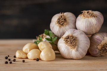 Garlic with peeled cloves, green sprig and whole black pepper on a wooden board with dark blurred background.