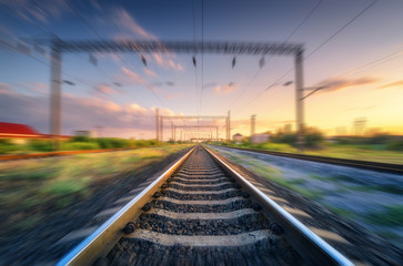 Fototapeta na wymiar Railroad and beautiful sky at sunset with motion blur effect in summer. Industrial landscape with railway station and blurred background with colorful sky. Railway platform in speed motion. Sleepers