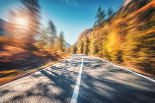 Mountain road in autumn forest at sunset with motion blur effect. Asphalt  road and blurred background with orange trees, blue sky with sun in fall.  Fast driving. Beautiful highway. Transportation Stock Photo |