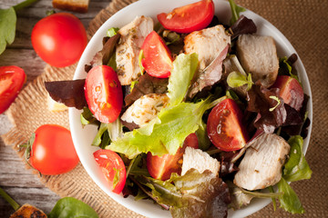 Fresh salad made of tomato, ruccola, chicken breast, eggs, arugula, crackers and spices. Caesar salad in a white, transparent bowl on wooden background