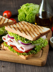 Close-up photo of a club sandwich. Sandwich with meat, prosciutto, salami, salad, vegetables, lettuce, tomato, onion and mustard on a fresh sliced rye bread on wooden background. Olives background.