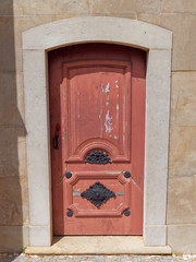 A wooden Door with faded and Peeling red paint, with highly decorated Metal Door Furniture set into a wall in the gardens of the Estoi Palace in Portugal.