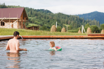 Dad with daughter swims in outdoor pool on mountains background.