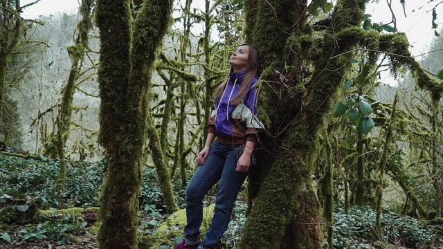 A girl stands leaning against a moss-covered tree in a mysterious forest