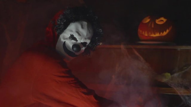 Unrecognizable man with scary clown halloween mask playing piano with mystic fog and pumpkin in the background. Scary halloween party performance at night