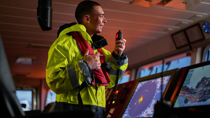 Navigator. pilot, captain as pat of ship crew performing daily duties with VHF radio, binoculars on board of modern ship with high quality navigation equipment on the bridge on sunrise.