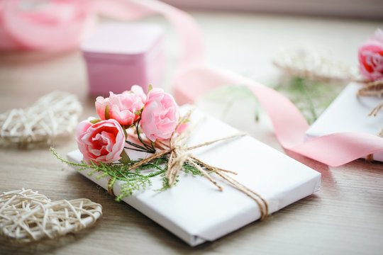 Handmade present box wrapped in white paper with branch of roses. Satin pink ribbon and wooden heart on the background.