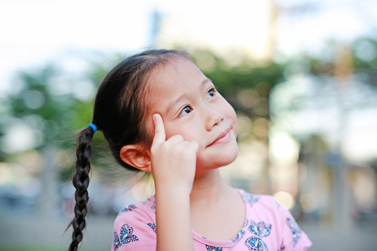 Portrait of happy little Asian child in garden with thinking and looking up. Close-up smiling kid girl with forefinger point on head in summer park.