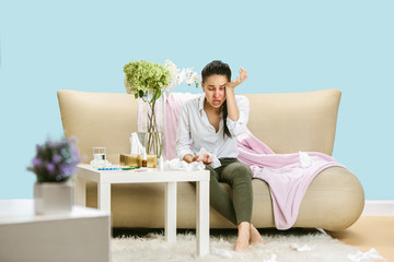 Young woman suffering from hausehold dust or seasonal allergy. Sneezing in the napkin and sitting surrounded by used napkins on the floor and sofa. Taking medicines with no result. Healthcare concept.