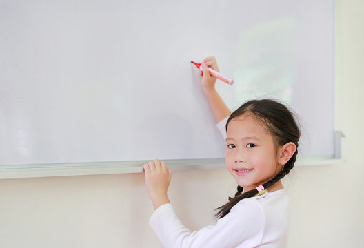 Portrait Of Happy Little Asian Child Girl Or Schoolgirl Writing Something On Whiteboard With A Marker And Looking At Camera In The Classroom. White Board With Copy Space For Text.